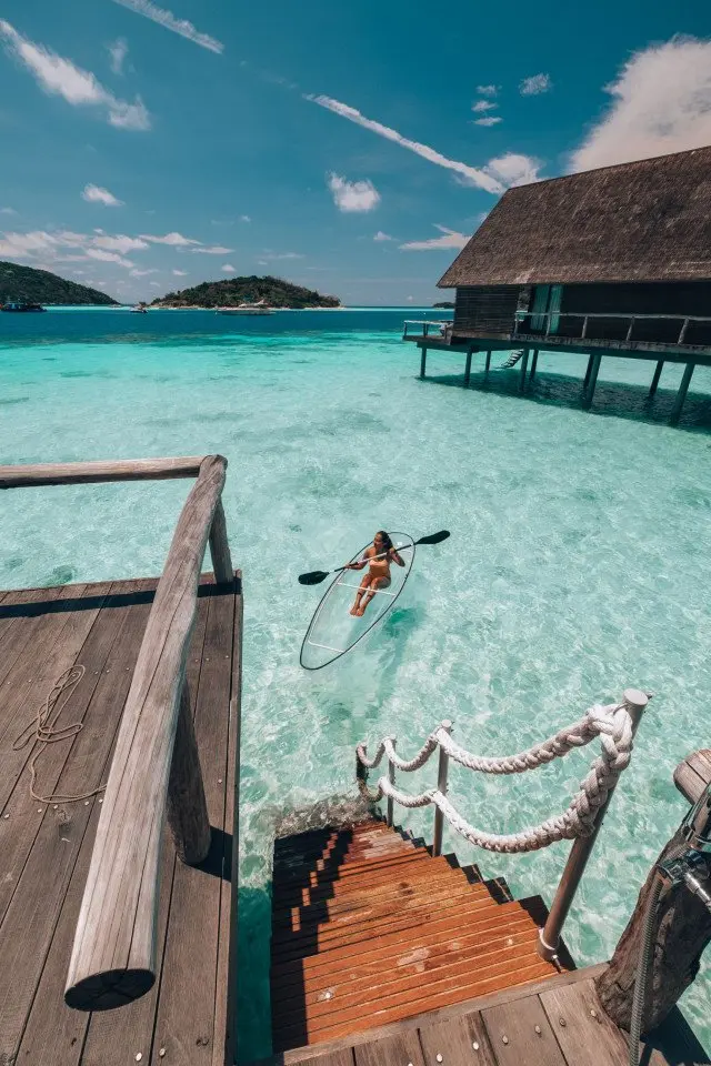 woman_clear_kayaking_towards_overwater_suite_wooden_steps_with_muerba_background